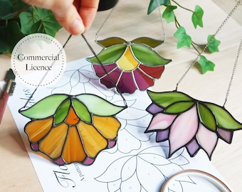 May include: Three stained glass flower suncatchers hanging from chains. The flowers are in shades of purple, pink, and yellow with green leaves. The suncatchers are hanging over a sheet of paper with a flower pattern.
