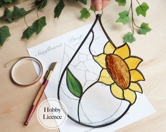 May include: A stained glass sunflower drop with a green leaf, hanging from a black metal frame. The glass pieces are yellow, brown, and green. The image also shows a pattern, a copper tape roll, and a glass cutter.