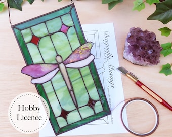 May include: A stained glass dragonfly window hanging with a green, pink, and white dragonfly design. The window is on a wooden surface with a sheet of paper with the text "Dragonfly Musings" and a hobby license.