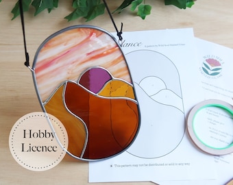 May include: A stained glass suncatcher with a hobby license sticker. The suncatcher is oval shaped and features a sunset scene with orange, yellow, and purple glass. The pattern is printed on a white sheet of paper with the text "Wild Soul Stained Glass" and "This pattern may not be distributed or sold in any way."