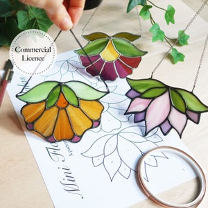 Puede incluir: Tres atrapasoles de flores de vidrio coloreado que cuelgan de cadenas. Las flores son en tonos de morado, rosa y amarillo con hojas verdes. Los atrapasoles cuelgan sobre una hoja de papel con un patrón de flores.