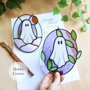 May include: Two oval stained glass suncatchers featuring white ghosts. One ghost is set against a pale pink and blue background with a small orange circle, while the other is surrounded by green glass leaves on a purple background with black solder lines.