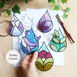 Puede incluir: Un conjunto de adornos de lágrima de vidrieras de colores con diseños florales y abstractos. Los adornos presentan tonos de morado, azul, verde y amarillo, con un borde de metal plateado. Una mano sostiene uno de los adornos. El texto "Hobby Licence" es visible.