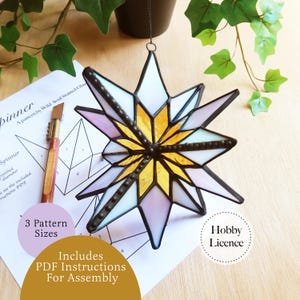 May include: A multi-pointed star ornament crafted from stained glass with white, yellow, and light purple panels joined by dark metal solder lines. The star is displayed alongside a printed paper pattern sheet and a glass cutting tool.