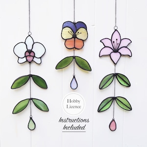 Op de afbeelding: Drie glas-in-lood bloemornamenten die aan zwarte kettingen hangen. De bloemen zijn een orchidee, een viooltje en een lelie, elk met groene bladeren en een druppelvormig accent. De tekst "Hobby Licence" en "Instructions Included" is ook zichtbaar.