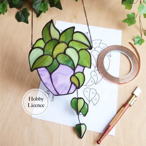 May include: Stained glass hanging planter with green and yellow leaves in a purple pot. Includes a pattern, copper tape, and a glass cutter. The Hobby Licence logo is visible.