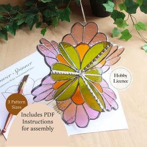 May include: A stained glass flower spinner featuring layered petals in shades of pink, orange, and lime green, joined by a central metal structure. The piece hangs from a silver chain and is displayed over a paper pattern labeled Flower Spinner.