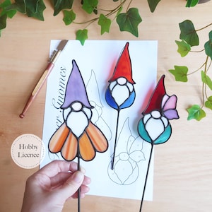 May include: Three colorful stained glass garden gnomes with black stems. One gnome has a purple hat and orange skirt, another has a red hat and blue body, and the third has a red hat and pink flower. The gnomes are on a white surface.