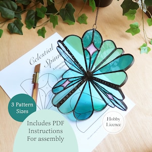 May include: A stained glass celestial spinner featuring teal and light blue petals with small pink diamond accents, joined by dark metal solder lines. The spinner rests on a printed paper pattern labeled Celestial Spinner with instructions.