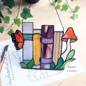 Peut inclure: Vitrail représentant une pile de livres, un papillon et un champignon. L'œuvre utilise des couleurs vives comme l'orange, le violet, le jaune et le vert. Une chaîne noire permet de l'accrocher.