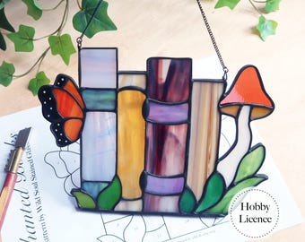Puede incluir: Vidriera que representa una pila de libros, una mariposa y una seta. La obra utiliza colores como naranja, morado, amarillo y verde. Tiene una cadena negra para colgar. Diseño inspirado en la naturaleza.