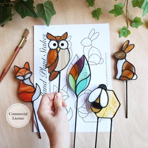 May include: A collection of stained glass plant stakes featuring a fox, owl, leaf, bee, and rabbit. The fox is orange and white, the owl is brown and beige, the leaf is green and purple, the bee is yellow and white, and the rabbit is brown and white.