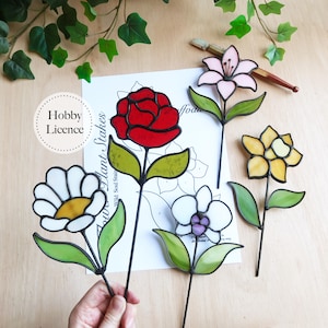 Peut inclure: Plusieurs tuteurs de plantes en forme de fleurs en vitrail coloré. On distingue une rose rouge, une marguerite blanche, un lis rose, une fleur jaune et une orchidée blanche et violette. Sur une surface en bois.