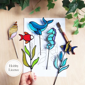 以下が含まれることがあります： ステンドグラスで作られたカラフルな海の生き物と植物のステークスのコレクション。クジラ、タツノオトシゴ、魚、貝殻、水生植物が含まれています。各作品は、庭や屋内のディスプレイ用の黒い金属製のステークに取り付けられています。