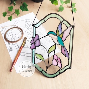 Op de afbeelding: Gebrandschilderd glazen kolibrie paneel met een groen, roze en turquoise ontwerp. Het paneel is zwart omlijst en heeft een ophangketting. De afbeelding toont ook een patroon, koperband en een glassnijder.