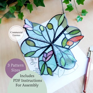 May include: A stained glass flower ornament with light blue, green, pink, and orange glass pieces held by dark metal solder lines. It rests on a printed paper pattern sheet labeled with text including Commercial Licence and 3 Pattern Sizes.