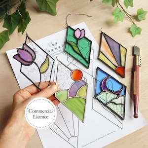 May include: Five stained glass diamond-shaped window hangings with black outlines. The designs include a purple tulip, a sunrise, a landscape, a crescent moon, and a pink tulip. The hangings are on a white sheet of paper with a printed pattern and the words "Mini Diamonds" and "A pattern by Soul Glass Studio".