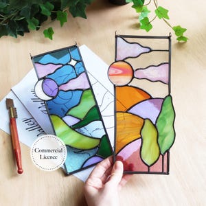 May include: Two stained glass panels depicting landscapes with a sun and moon. One panel features a blue sky with a moon, the other a sunset with orange and pink hues. The panels are rectangular with black metal frames.