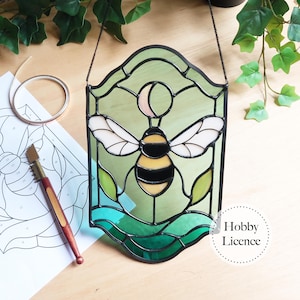 May include: Stained glass panel with a bee design, incorporating yellow, black, and white elements. The panel has a green and blue backdrop with leaf motifs. It is suspended by a chain. Crafting supplies are also visible in the image.
