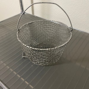 May include: A silver wire mesh basket with a handle. The basket is empty and sitting on a gray metal surface.