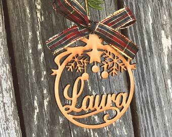 May include: A wooden Christmas ornament with the name "Laura" cut out in a cursive font. The ornament is round with snowflake and star details, and is topped with a plaid ribbon. The ornament hangs from a green branch with red berries.