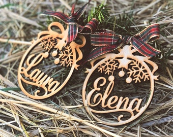 May include: Two wooden Christmas ornaments with the names "Laura" and "Elena" cut out. The ornaments are oval-shaped with snowflake and star designs. Each ornament has a red, green, and gold plaid ribbon and a small sprig of evergreen.