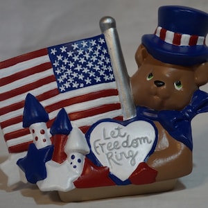 May include: A brown bear figurine wearing a blue top hat with red and white stripes, holding a white heart with the text "Let Freedom Ring" in silver lettering. The bear is holding an American flag with blue and white stars on a red and white striped background. The figurine is decorated with blue and white stars and red and white fireworks.