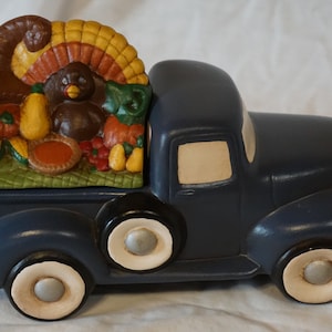 May include: A blue ceramic pickup truck with a white cab and a bed full of Thanksgiving-themed decorations. The decorations include a cornucopia, a turkey, pumpkins, and other fall harvest items.