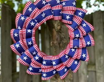 May include: A patriotic wreath made of blue ribbon with white stars and red and white striped edges. The wreath is hanging from a tree branch, perfect for Independence Day or other patriotic holidays.