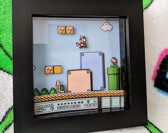 May include: A framed 3D diorama featuring a pixelated scene from the Super Mario Bros. video game. The scene includes Mario, question mark blocks, a Piranha Plant, and other iconic elements, all set against a blue sky background.