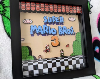 May include: Framed artwork featuring a pixelated scene from Super Mario Bros. 3. The artwork includes the game's title, characters Mario and Luigi, and a checkered foreground. The frame is black.