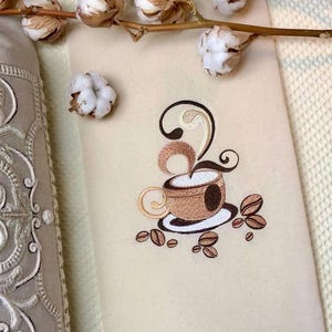 Puede incluir: Toalla de cocina bordada con un diseño de taza de café marrón y vapor. La taza está sobre un platillo blanco con granos de café marrones esparcidos alrededor.
