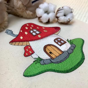 以下が含まれることがあります： 赤い帽子、白い壁、茶色のドアが付いたキノコの家が刺繍されたデザイン。家は緑の草と灰色の岩に囲まれています。