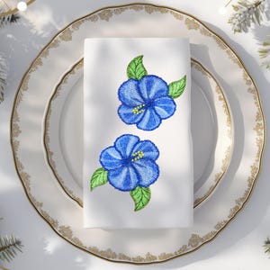 Op de afbeelding: Witte linnen servet geborduurd met twee blauwe hibiscusbloemen en groene bladeren. De servet is gevouwen en op een wit bord met een gouden rand geplaatst. Het bord maakt deel uit van een set, perfect voor een formeel diner.