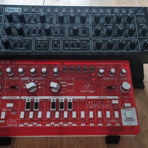 May include: A black Behringer Pro-1 analogue synthesiser with numerous knobs and switches sits behind a translucent red Behringer TD-3 bass line synthesiser featuring white knobs, a step sequencer, and various control buttons on its top panel.