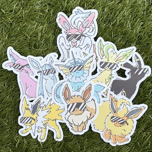 May include: A selection of cartoon stickers depicting various characters, all sporting sunglasses. The stickers are in shades of pink, blue, yellow, and brown, with black sunglasses. The stickers are arranged on a green surface.