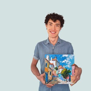 May include: A man in a blue shirt is holding a painting of a colorful cityscape with a blue sky and white clouds. The painting is framed and has a white border.