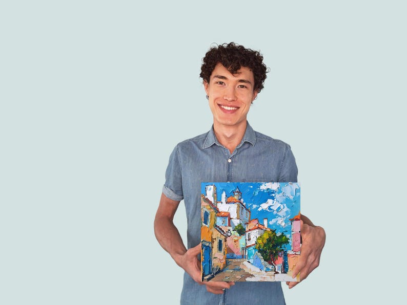May include: A man in a blue shirt is holding a painting of a colorful cityscape with a blue sky and white clouds. The painting is framed and has a white border.