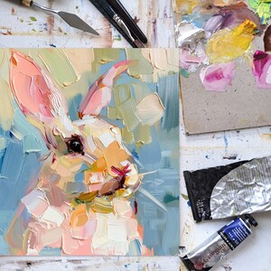 May include: A close-up painting of a white rabbit with pink ears and a blue background. The painting is done in a loose, impressionistic style with visible brushstrokes.