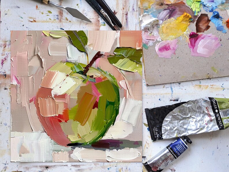 May include: A close-up of a painting of a green apple with pink and yellow highlights. The apple is painted in a loose, impressionistic style with visible brushstrokes. The background is a soft pink.