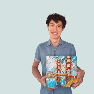 May include: A man wearing a blue shirt is holding a painting of the Golden Gate Bridge. The painting is done in a colourful, abstract style.