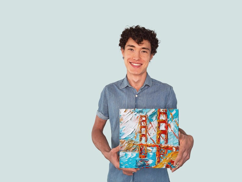 May include: A man wearing a blue shirt is holding a painting of the Golden Gate Bridge. The painting is done in a colourful, abstract style.