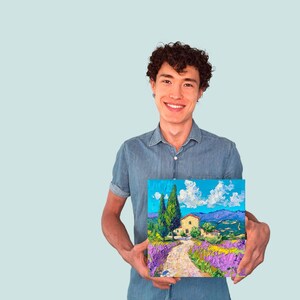 以下が含まれることがあります： 人が、家、木、ラベンダーの花が描かれた風景画を持っています。絵は明るくカラフルなスタイルです。