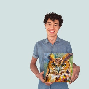 May include: A man is holding a painting of an owl with a colorful background. The owl has yellow eyes and brown feathers. The painting is framed and has a white border.