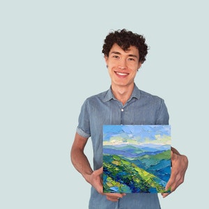 Op de afbeelding: Een man houdt een schilderij vast van een landschap met blauwe en groene heuvels en een blauwe lucht. Het schilderij is ingelijst en heeft een textuur.