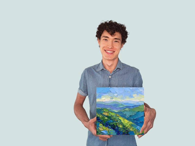 Op de afbeelding: Een man houdt een schilderij vast van een landschap met blauwe en groene heuvels en een blauwe lucht. Het schilderij is ingelijst en heeft een textuur.
