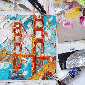 May include: A close-up of a painting depicting the Golden Gate Bridge in San Francisco. The painting is executed in a thick, impasto style using vibrant colours of red, orange, yellow, blue and white.