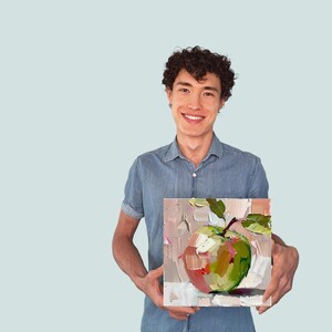 May include: A person holding a painting of a green apple with a pink blush on a white background.