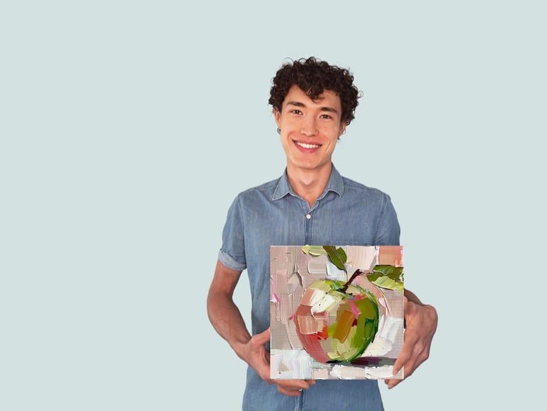 May include: A person holding a painting of a green apple with a pink blush on a white background.