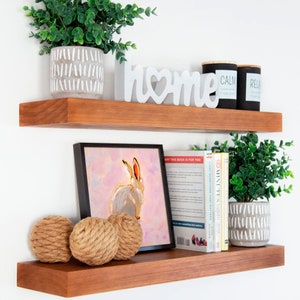 May include: Two wooden shelves with a white "home" sign, a painting of a white rabbit, a potted plant, and books. The shelves are mounted on a white wall.